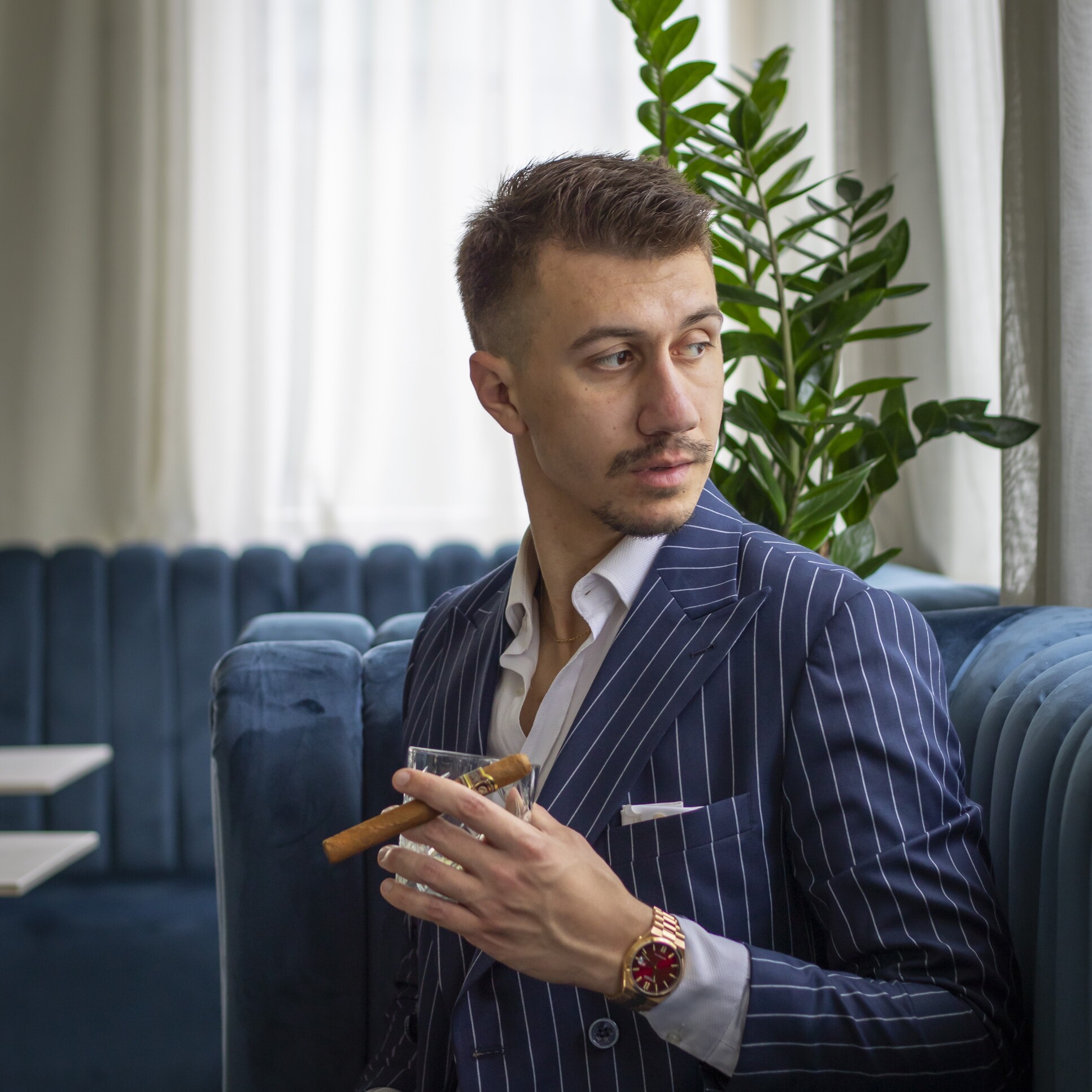 A man in a pinstripe suit holding a cigar and a glass, seated in a modern lounge with plants in the background, by Vojislav Zivkovic aka Vojke Photo